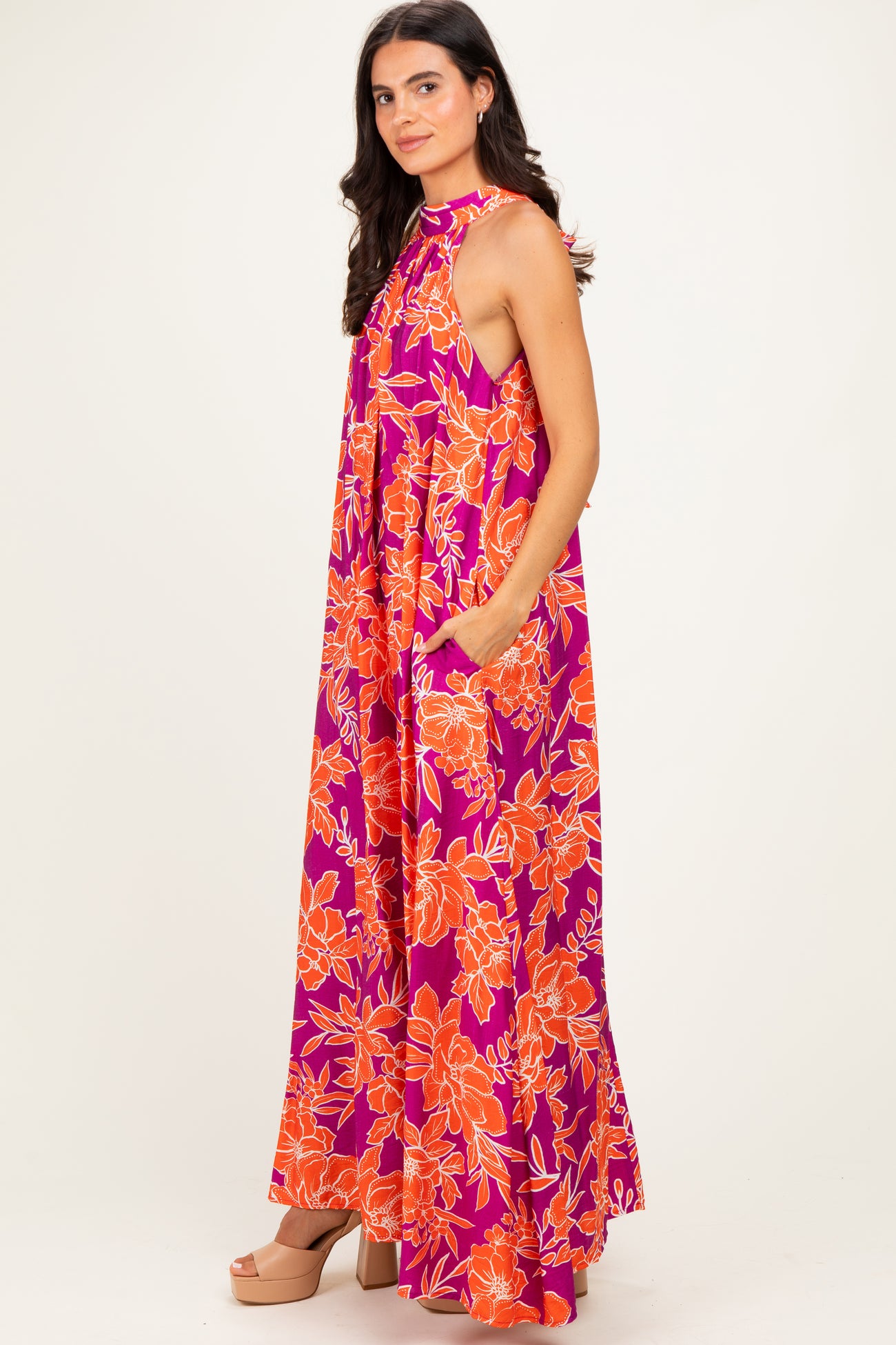 woman wearing a floral print halter neck maxi dress in bright pink and orange, featuring side pockets and a flowy silhouette