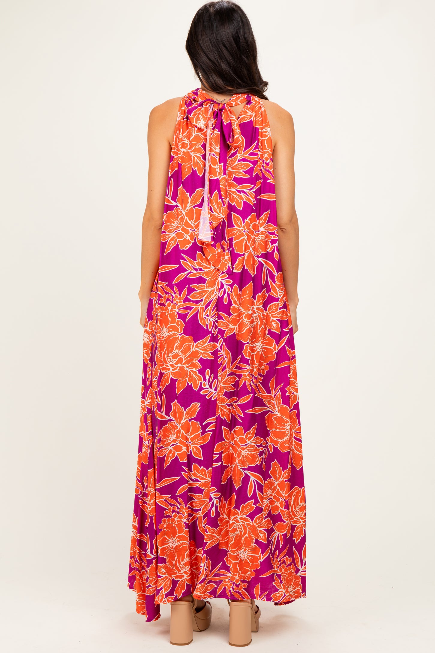 woman wearing a back view of a sleeveless purple maxi dress with orange floral print and a tie closure at the neck