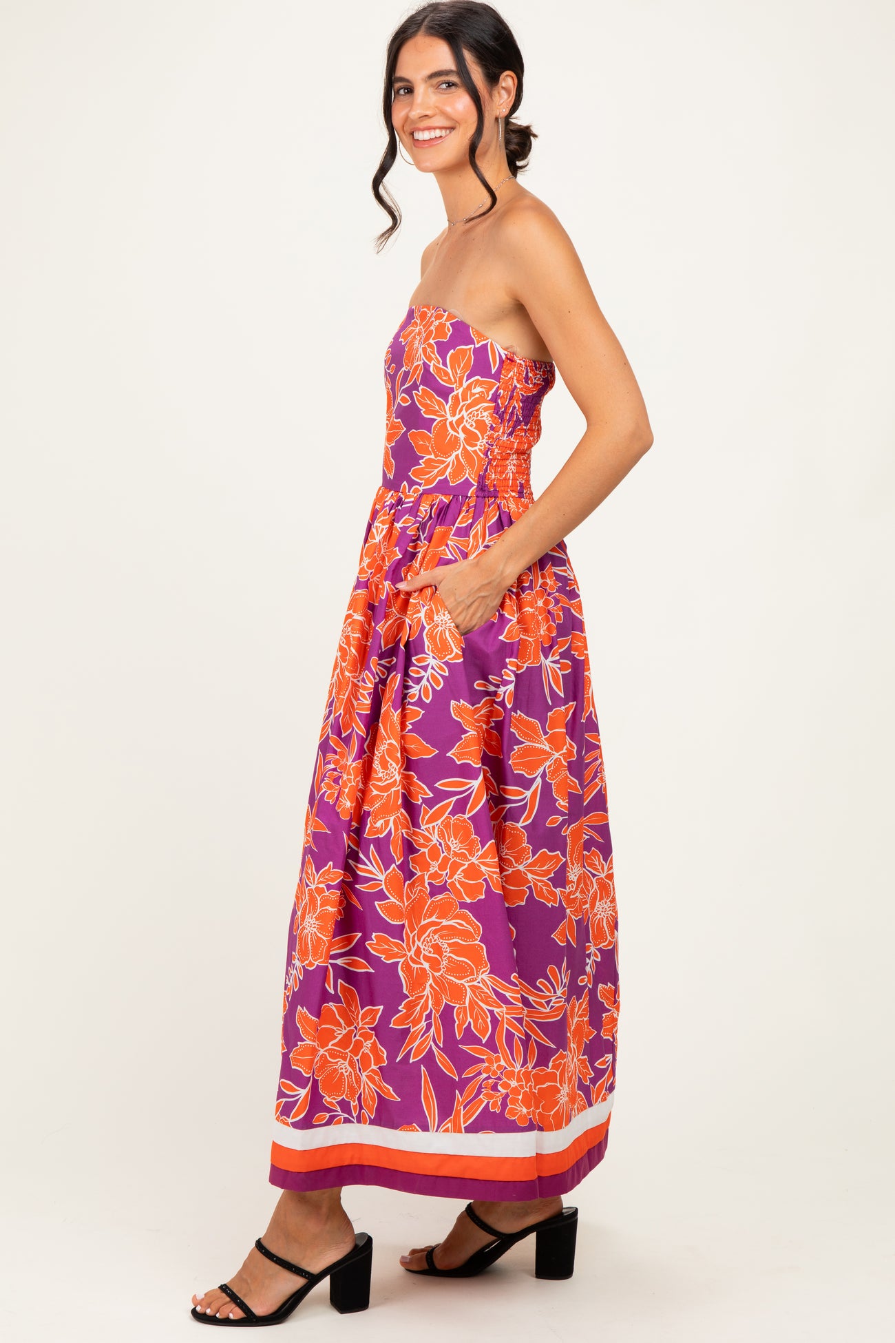 woman wearing a strapless purple floral maxi dress with orange details and pockets, standing sideways with a smile