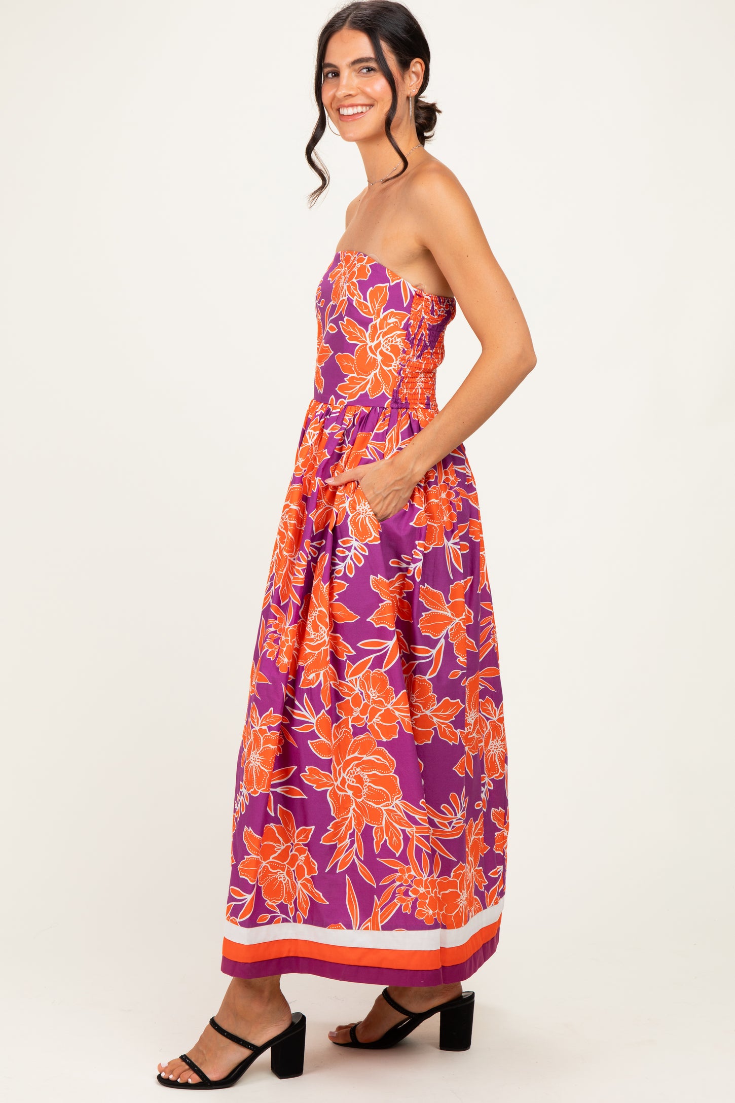 woman wearing a strapless purple floral maxi dress with orange details and pockets, standing sideways with a smile