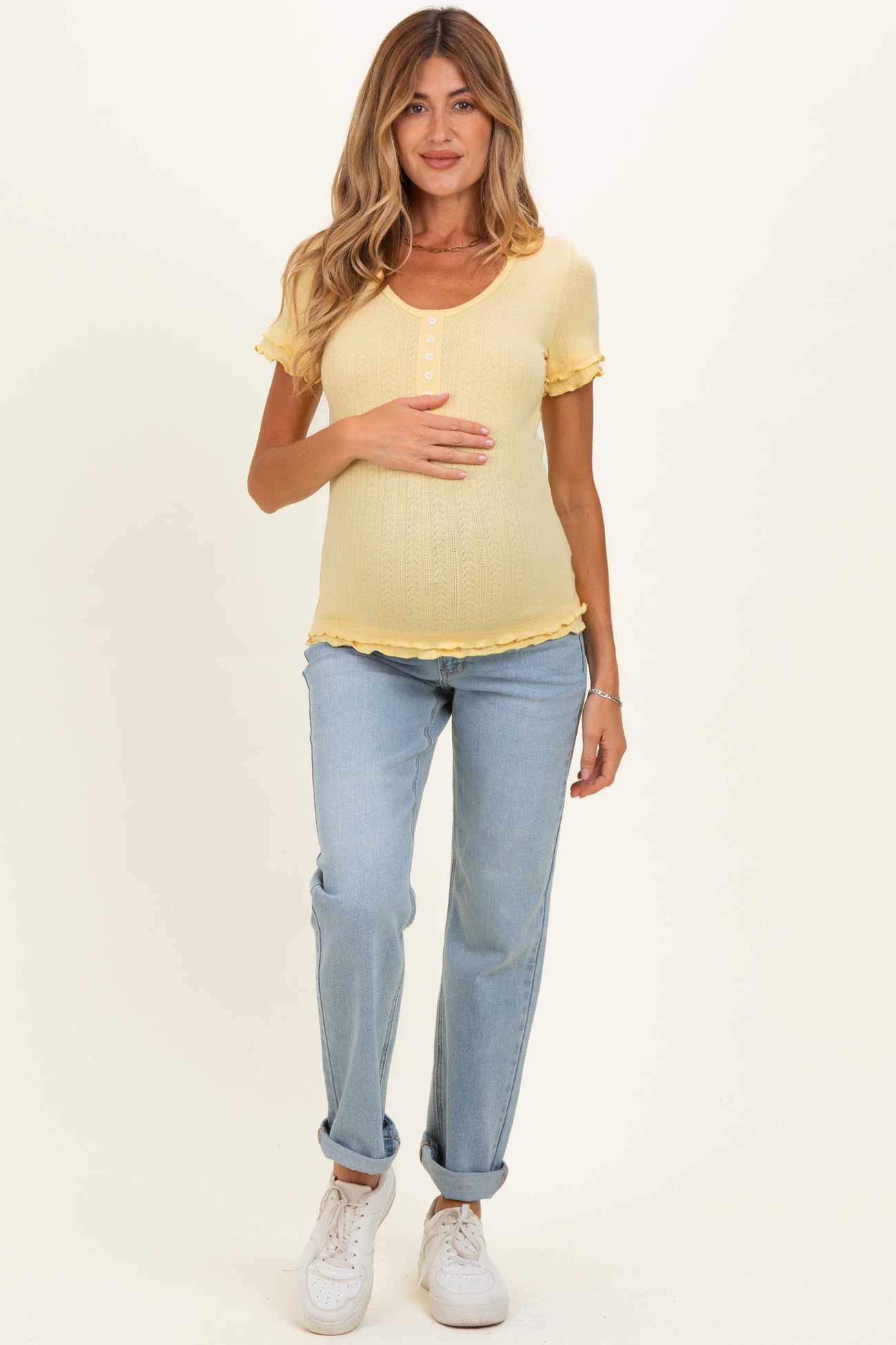 woman wearing a light yellow short sleeve maternity top with ruffle details and holding her baby bump, paired with light blue jeans