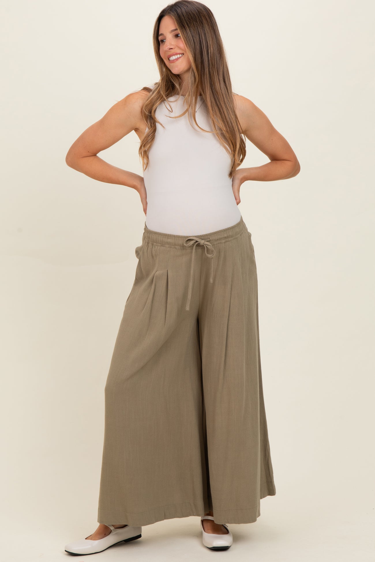 woman wearing olive green maternity wide-leg pants with a drawstring waist and a fitted white tank top, smiling with hands on hips