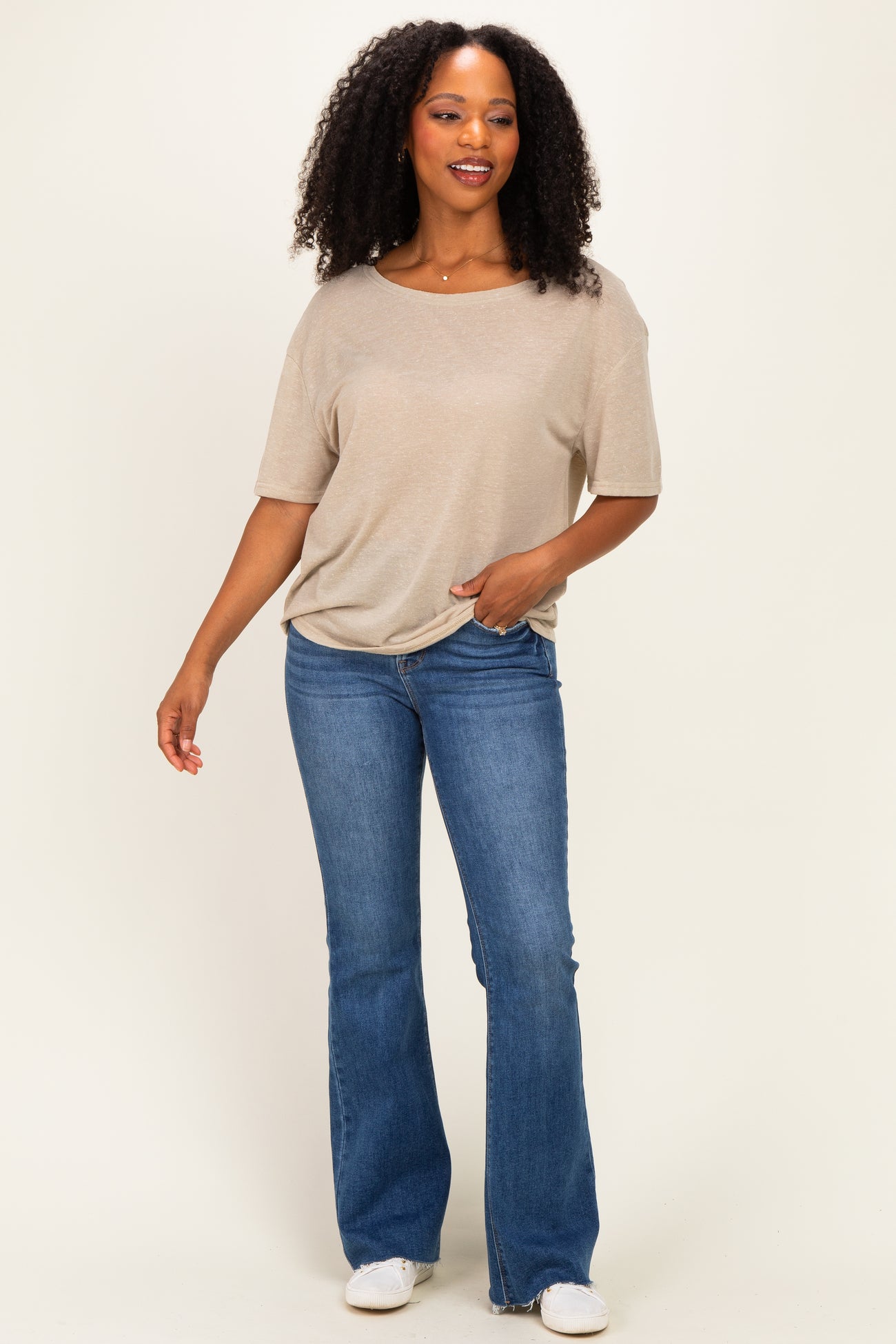 woman wearing a beige short sleeve top and blue flared jeans, standing and smiling against a neutral background