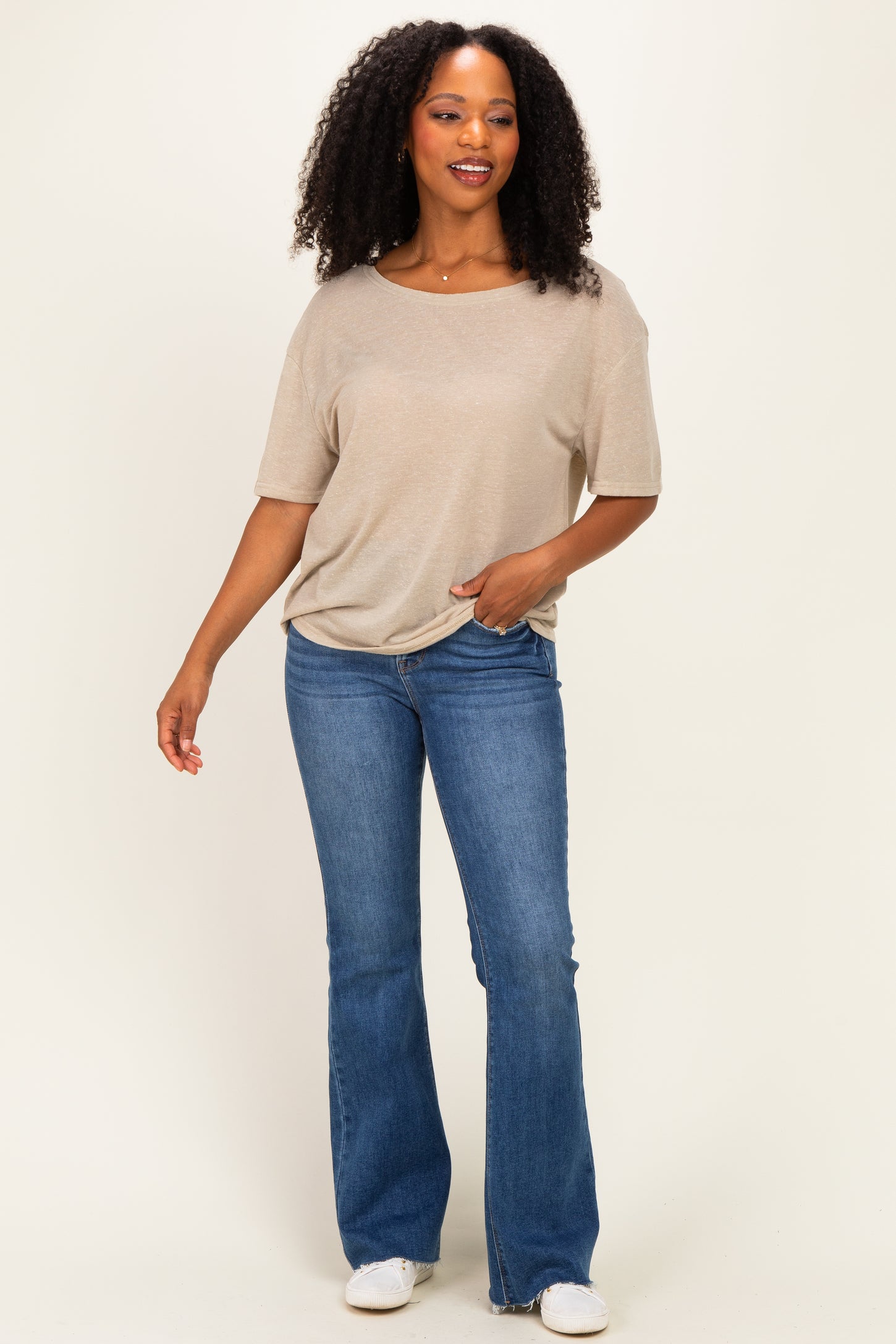 woman wearing a beige short sleeve top and blue flared jeans, standing and smiling against a neutral background