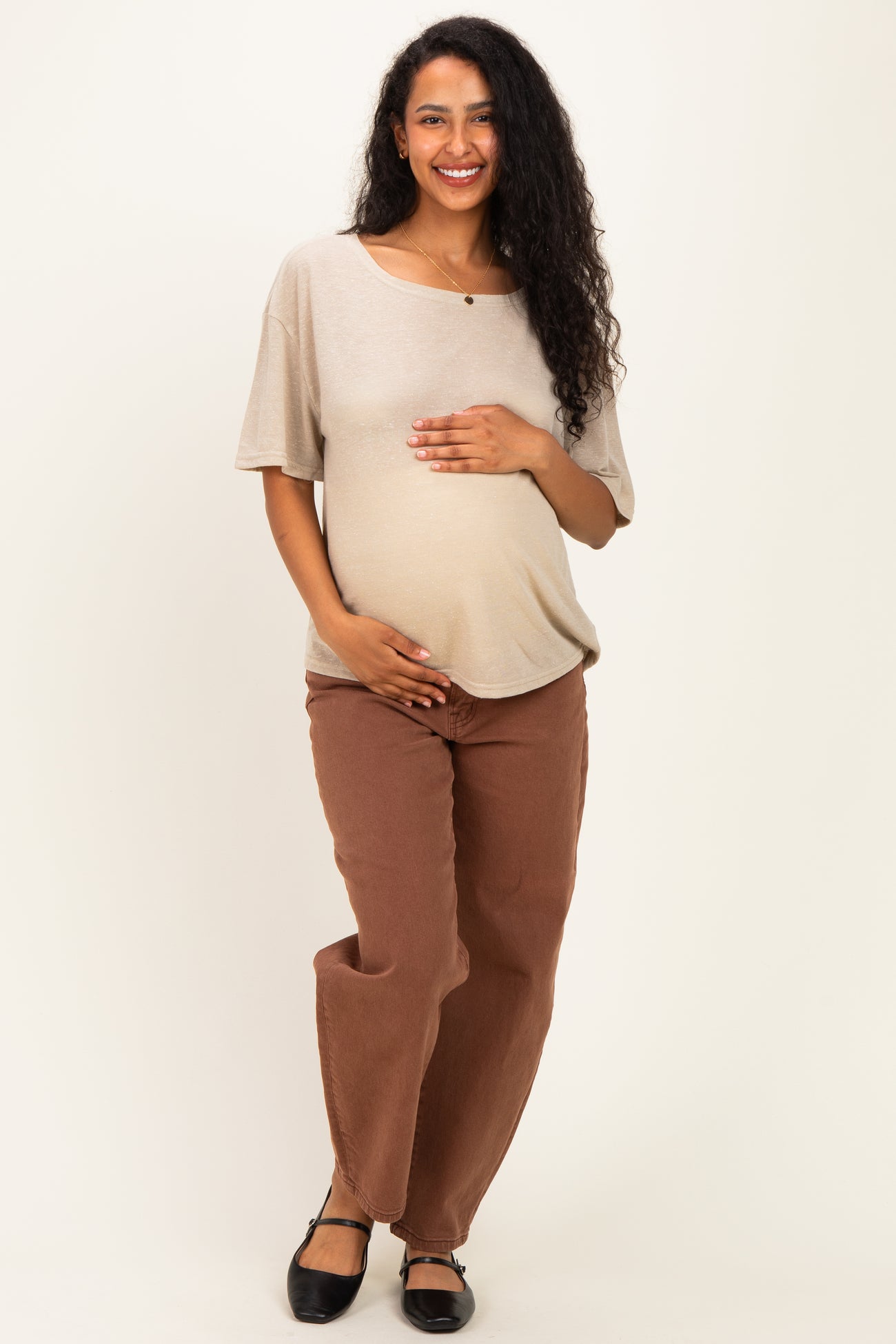woman wearing a beige short sleeve maternity top and brown maternity pants, gently holding her baby bump