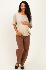 woman wearing a beige short sleeve maternity top and brown maternity pants, gently holding her baby bump