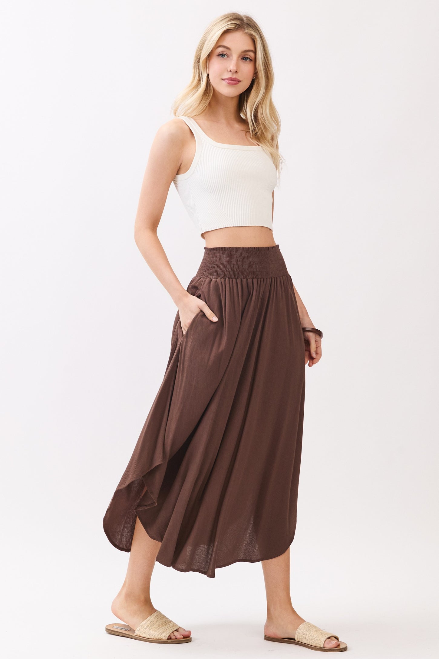 woman wearing a brown smocked waist maxi skirt and a white crop top, posing with one hand in the pocket