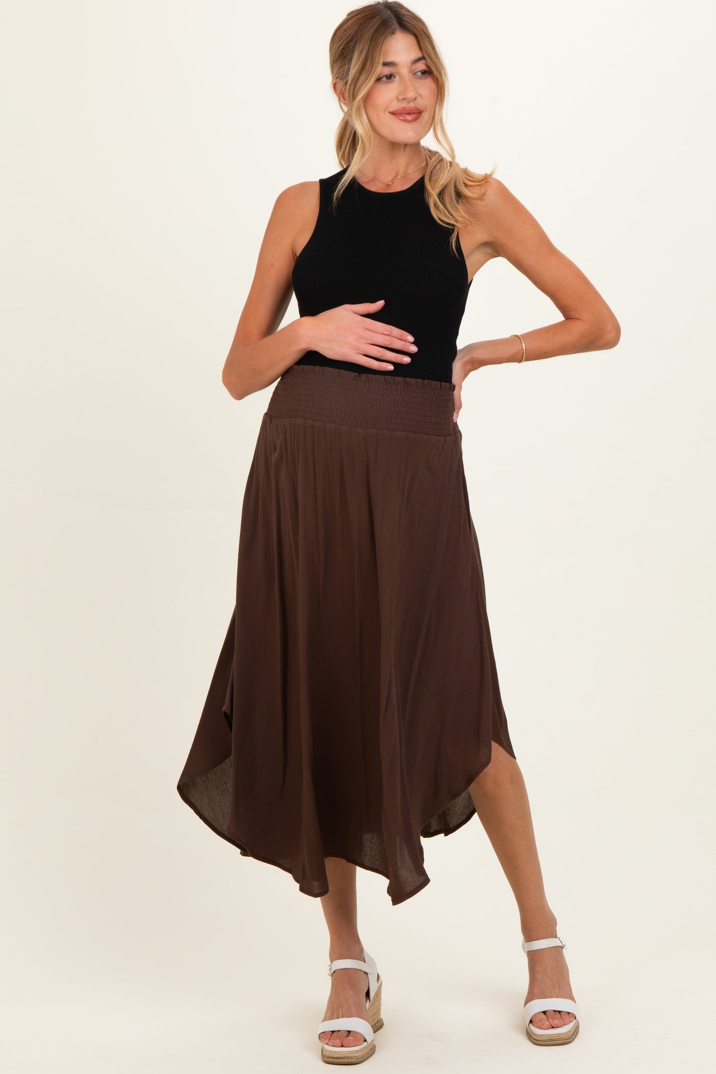 woman wearing a black sleeveless top and a brown asymmetrical maternity skirt, holding her baby bump