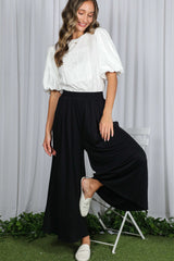 woman wearing a white puff sleeve blouse and black wide leg pants, posing with one foot raised on a chair