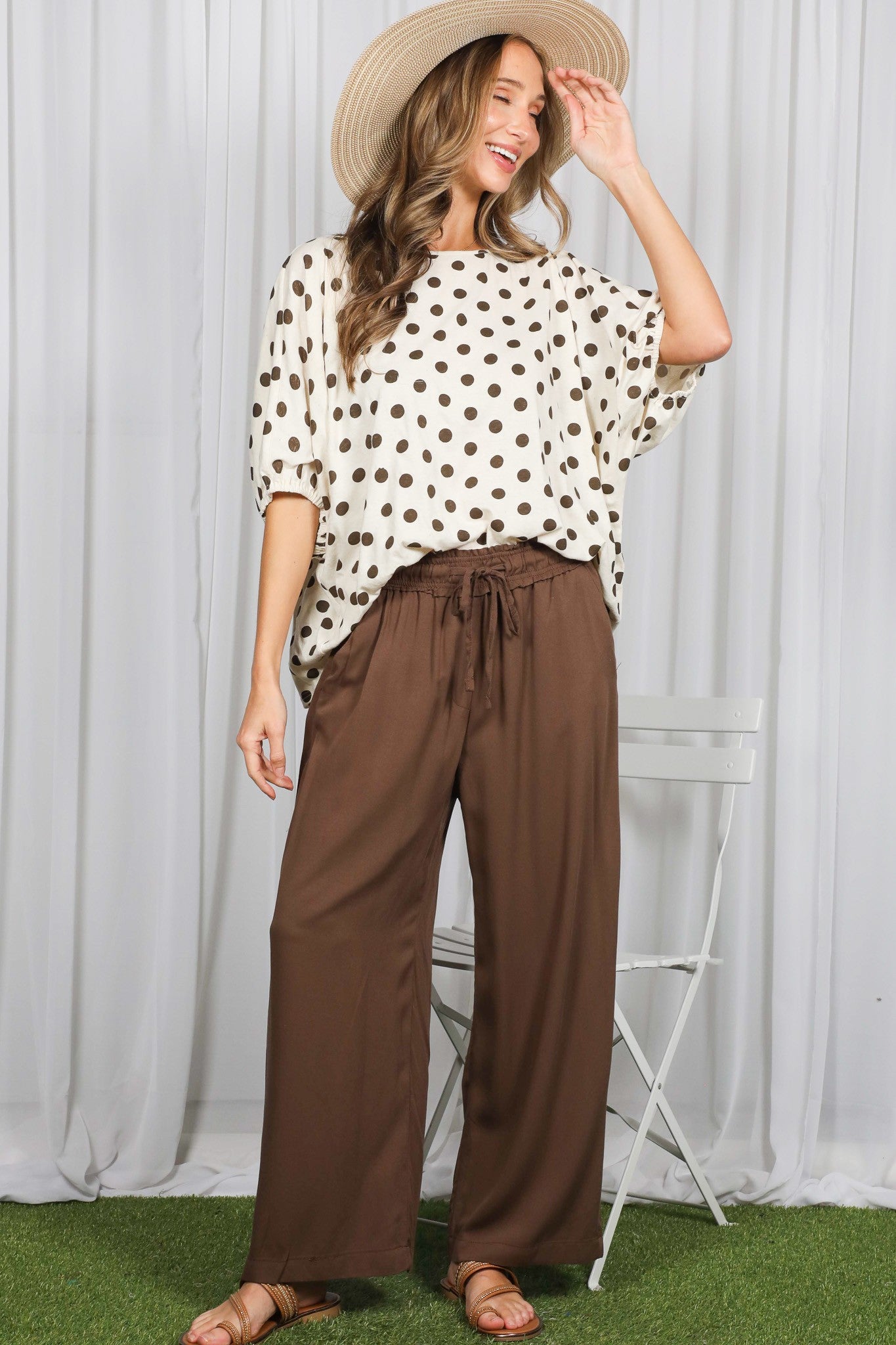 woman wearing a white polka dot oversized top and brown wide-leg pants, smiling and wearing a straw hat