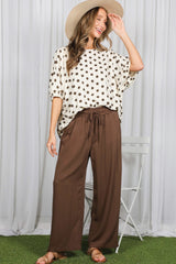 woman wearing a white polka dot oversized top and brown wide-leg pants, smiling and wearing a straw hat
