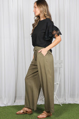 woman wearing a black top with lace ruffled sleeves and olive green wide-leg pants, standing against a white backdrop