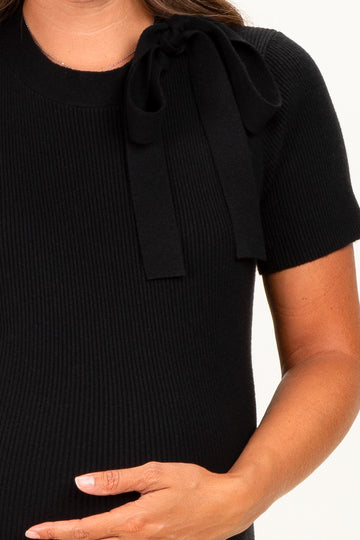 woman wearing a black short sleeve maternity knit top with a bow detail on the shoulder and holding her baby bump