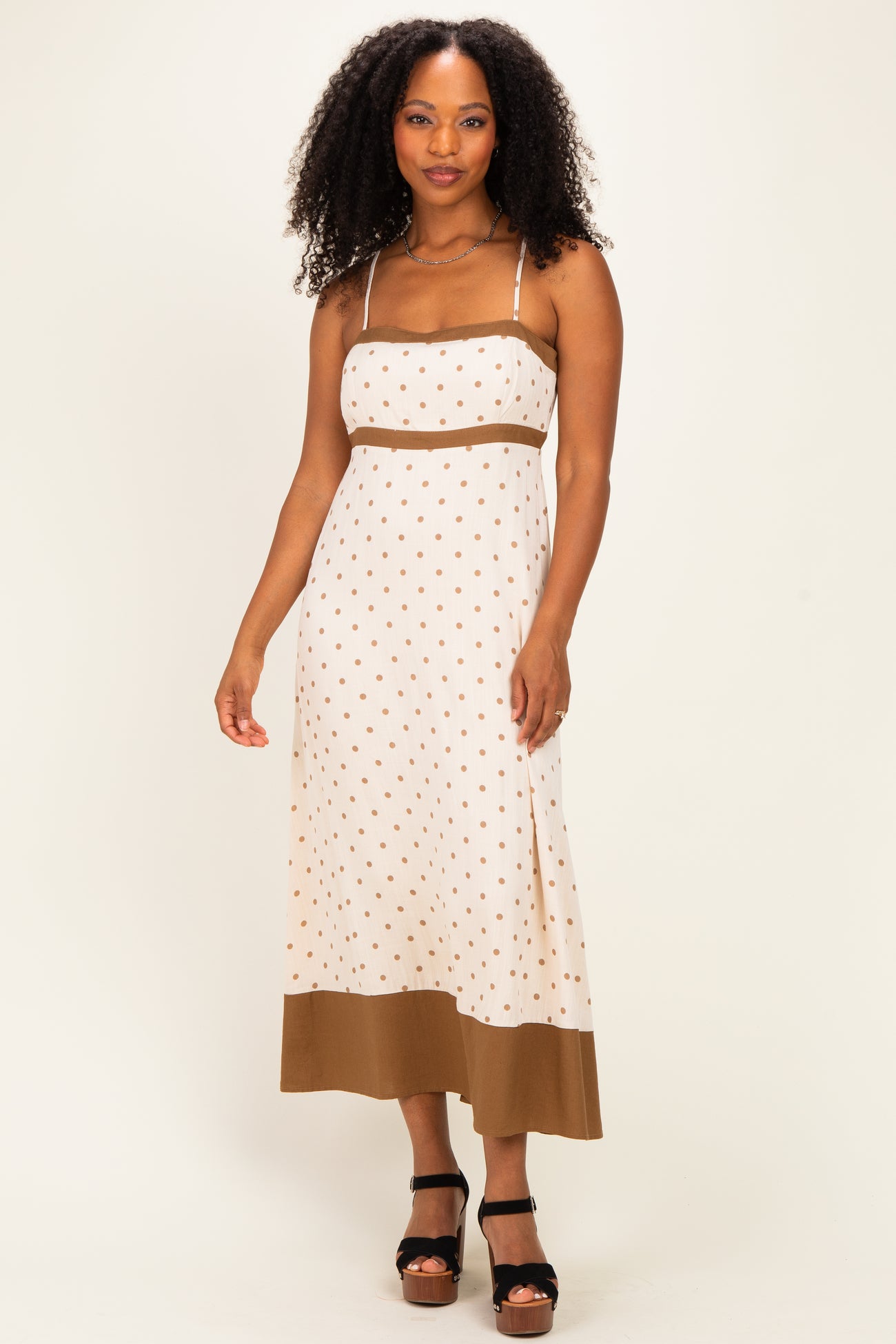 woman wearing a cream polka dot maxi dress with adjustable straps and a brown hem, standing in a pose
