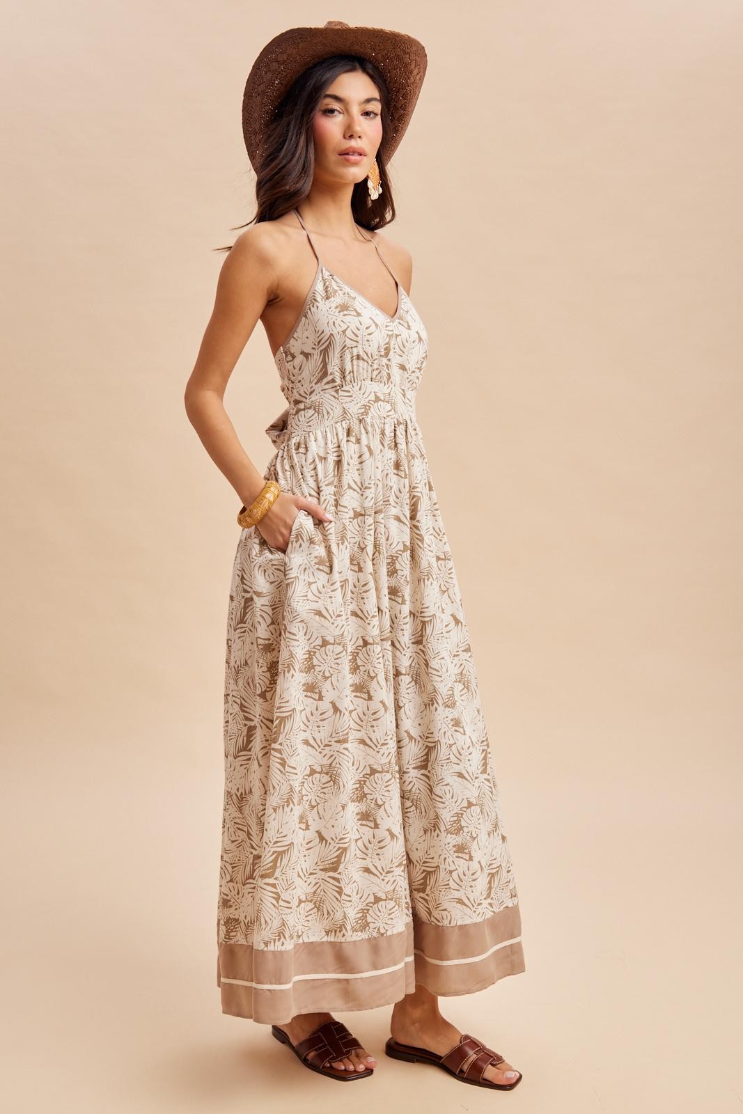 woman wearing a beige maxi dress with a floral print, thin straps, pockets, and a brown straw hat