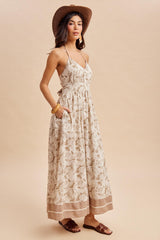 woman wearing a beige maxi dress with a floral print, thin straps, pockets, and a brown straw hat