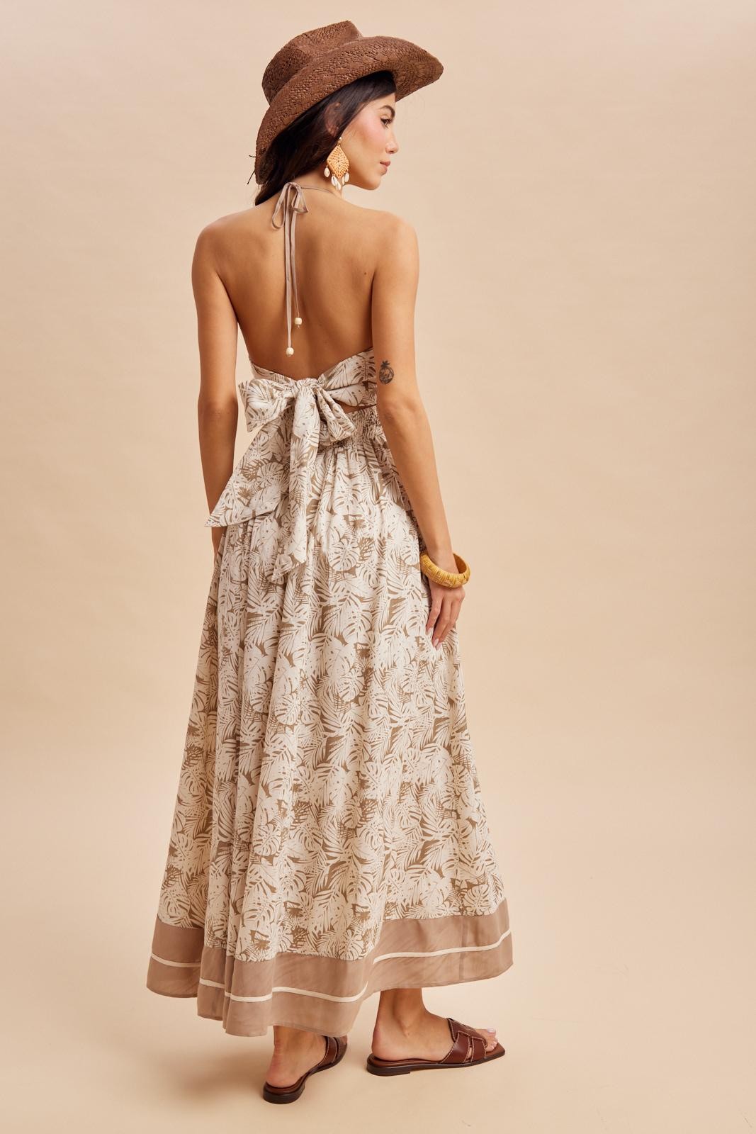 woman wearing a beige floral maxi dress with an open back and a large bow, styled with a brown hat and sandals