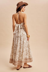 woman wearing a beige floral maxi dress with an open back and a large bow, styled with a brown hat and sandals