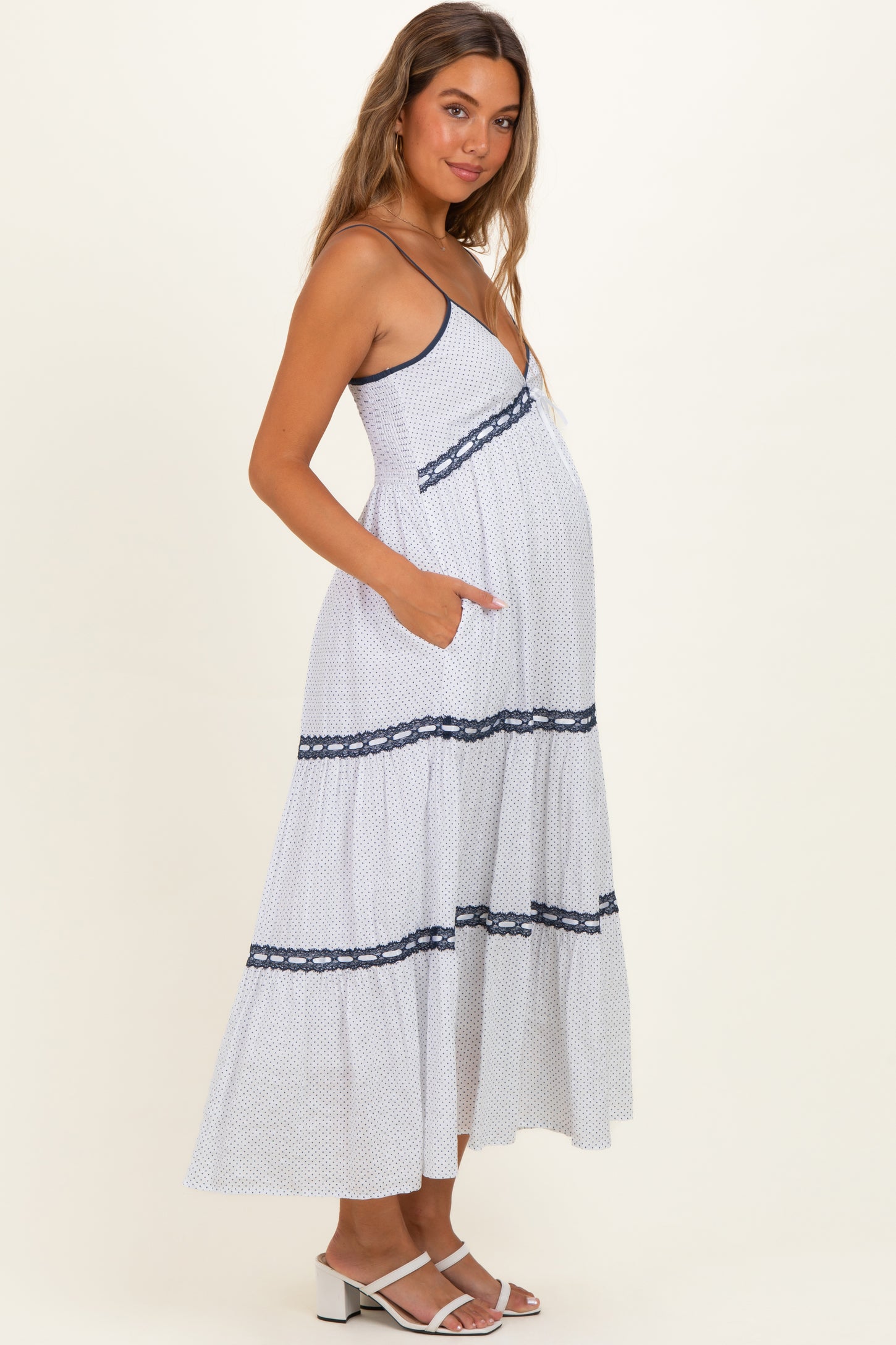 woman wearing a white and navy polka dot maternity maxi dress with spaghetti straps and pockets, showing her baby bump