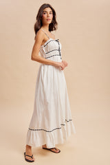 woman wearing a white maxi dress with black trim and a gathered bodice, standing against a neutral background