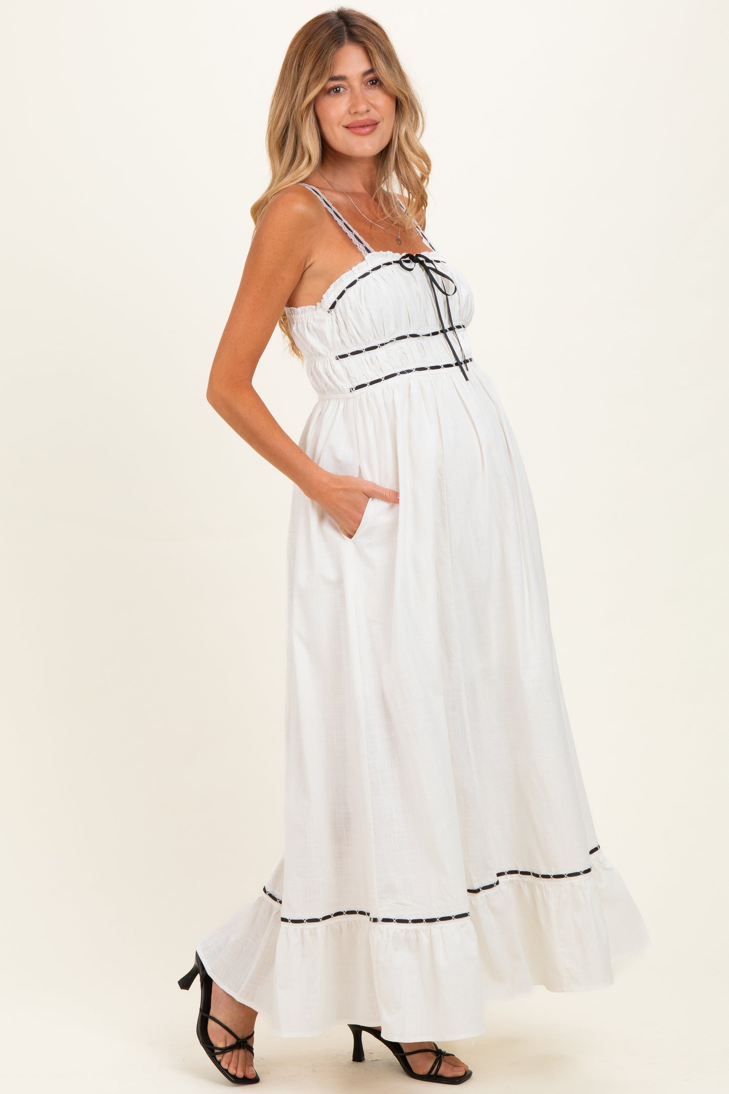 woman wearing a white maternity maxi dress with black trim, spaghetti straps, and pockets, showing her baby bump