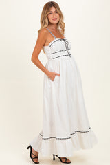 woman wearing a white maternity maxi dress with black trim, spaghetti straps, and pockets, showing her baby bump