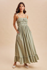 woman wearing a sage green maxi dress with adjustable straps, smocked bodice, and ruffled hem, styled with pockets