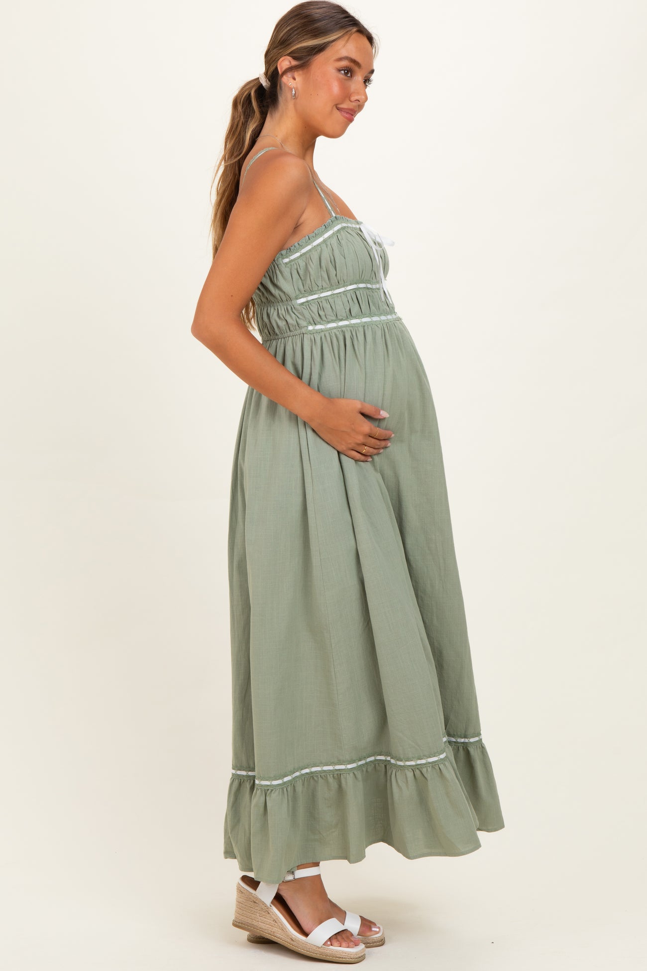 woman wearing a sage green maternity maxi dress with adjustable straps and ruffle hem, cradling her baby bump