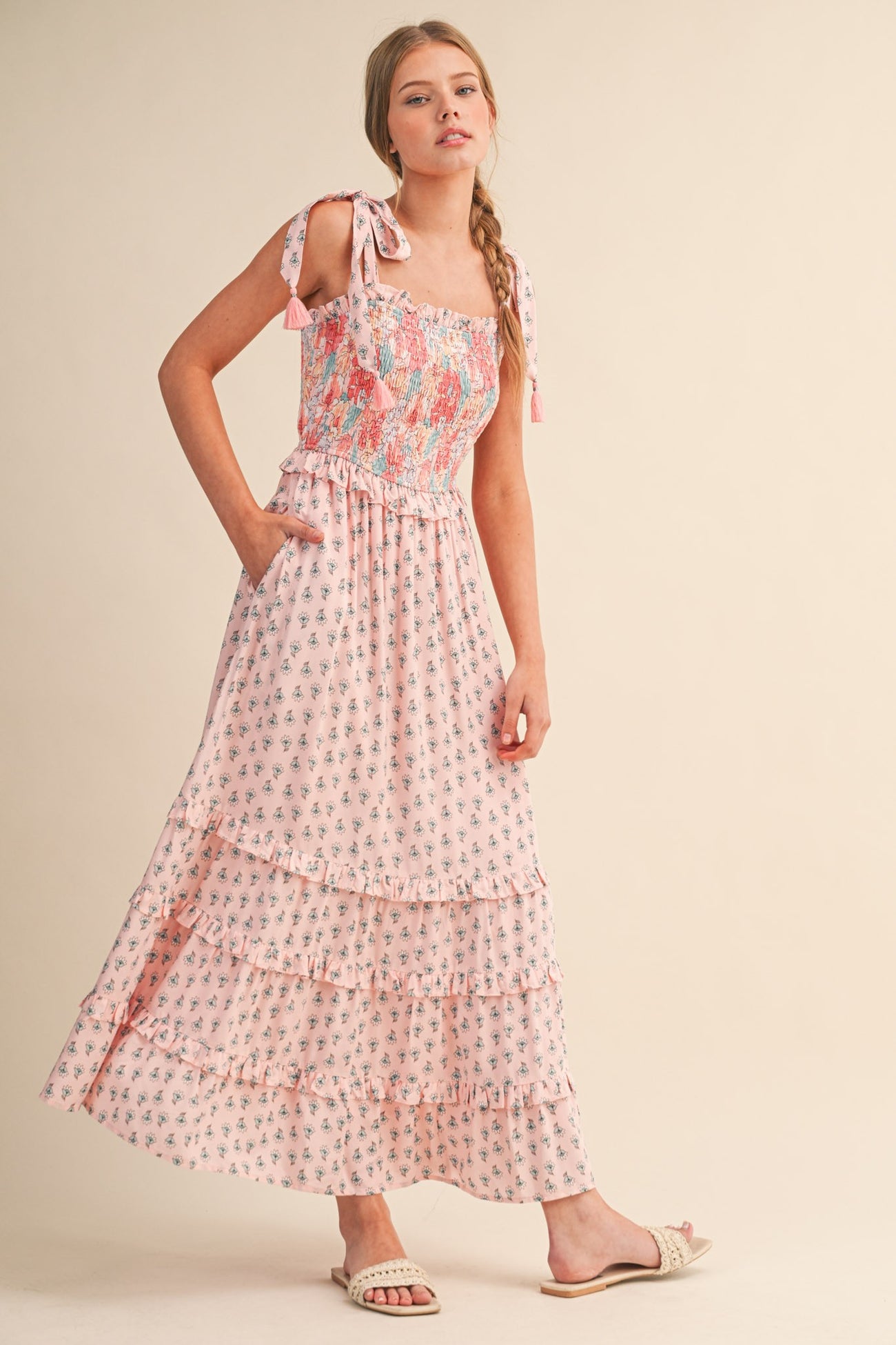 woman wearing a pink floral maxi dress with tie straps and a tiered skirt, standing with one hand in her pocket