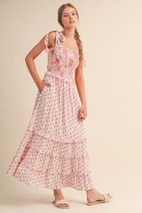 woman wearing a pink floral maxi dress with tie straps and a tiered skirt, standing with one hand in her pocket