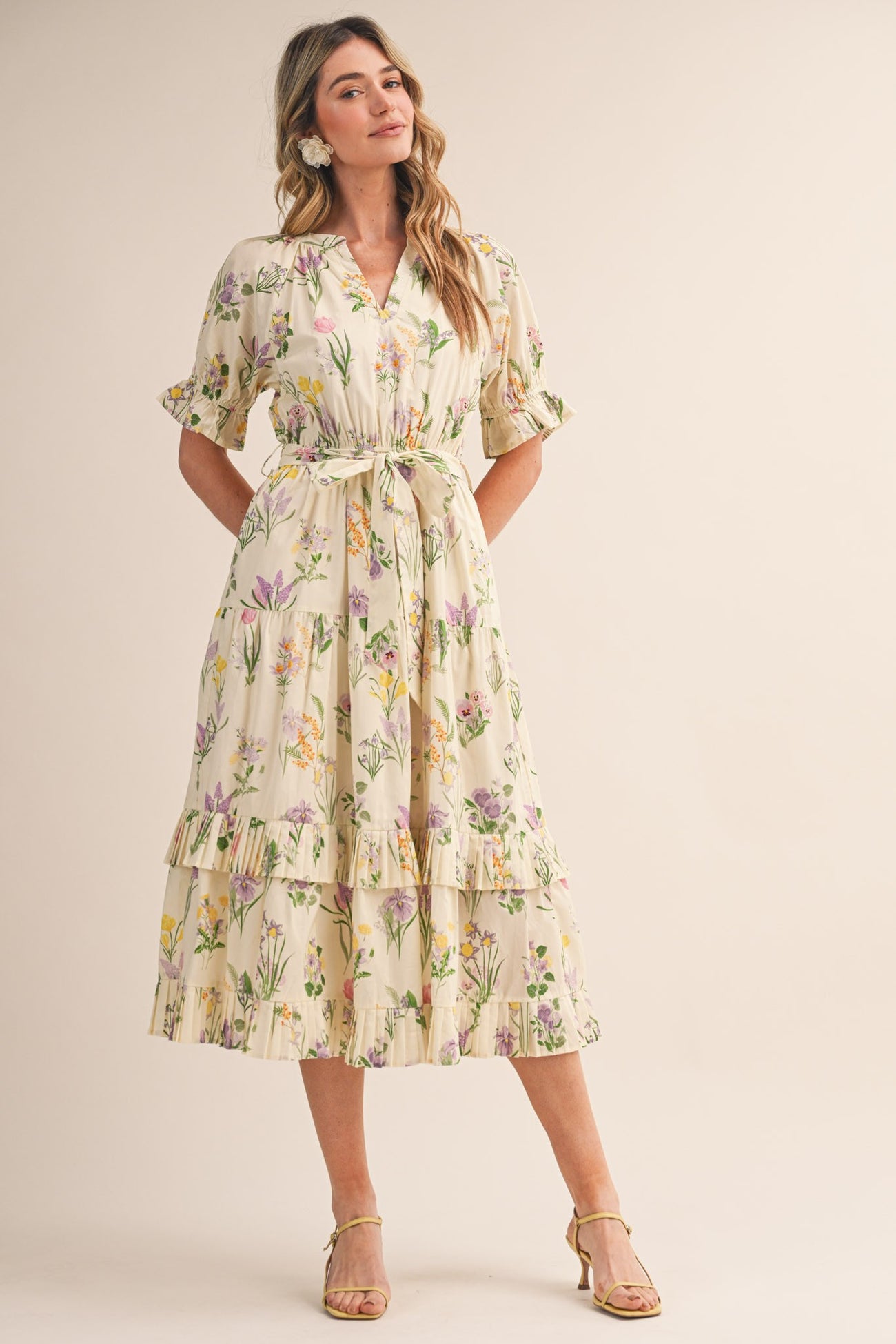 woman wearing a cream floral midi dress with tiered layers and a tie belt, standing on a neutral background
