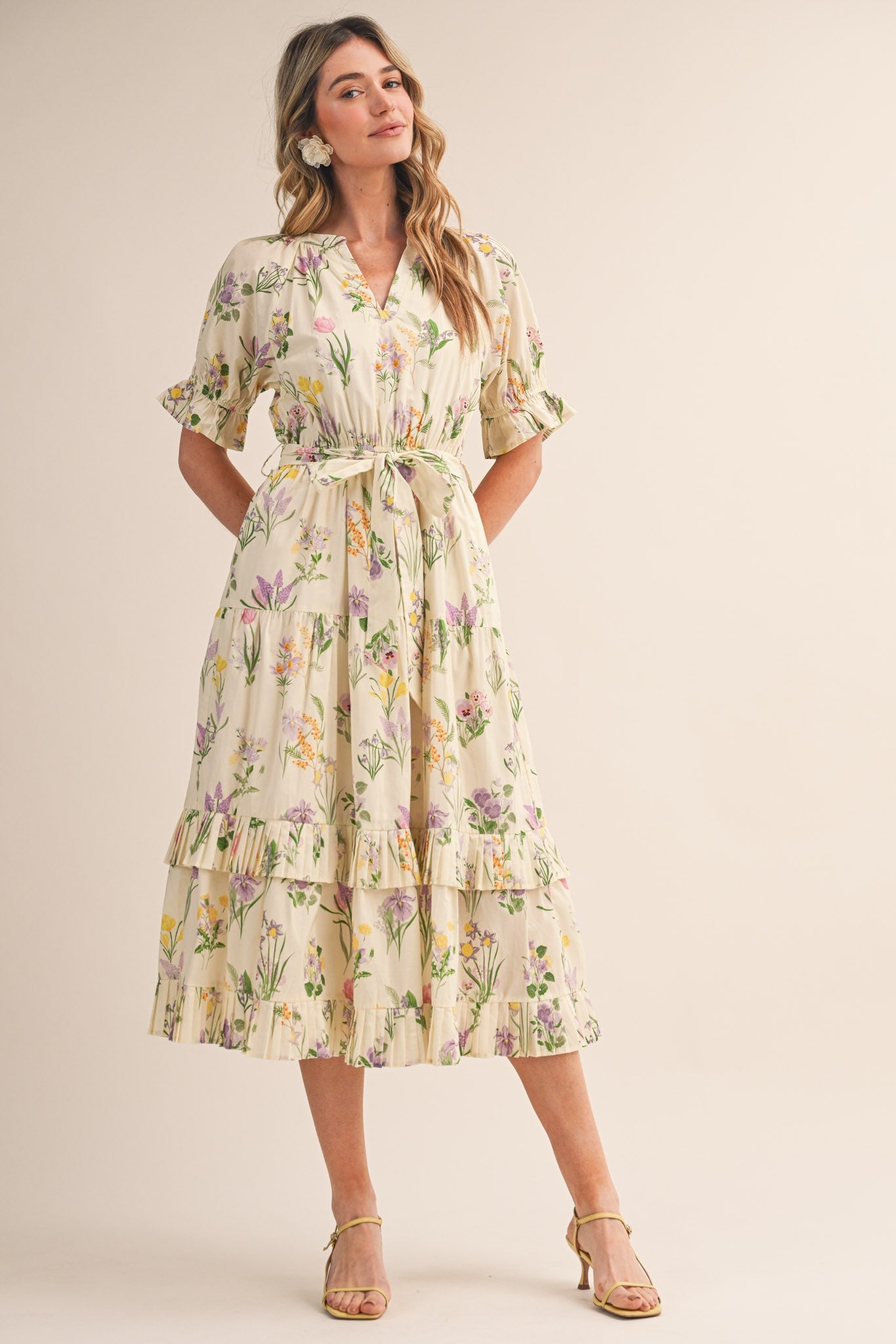 woman wearing a cream floral midi dress with tiered layers and a tie belt, standing on a neutral background