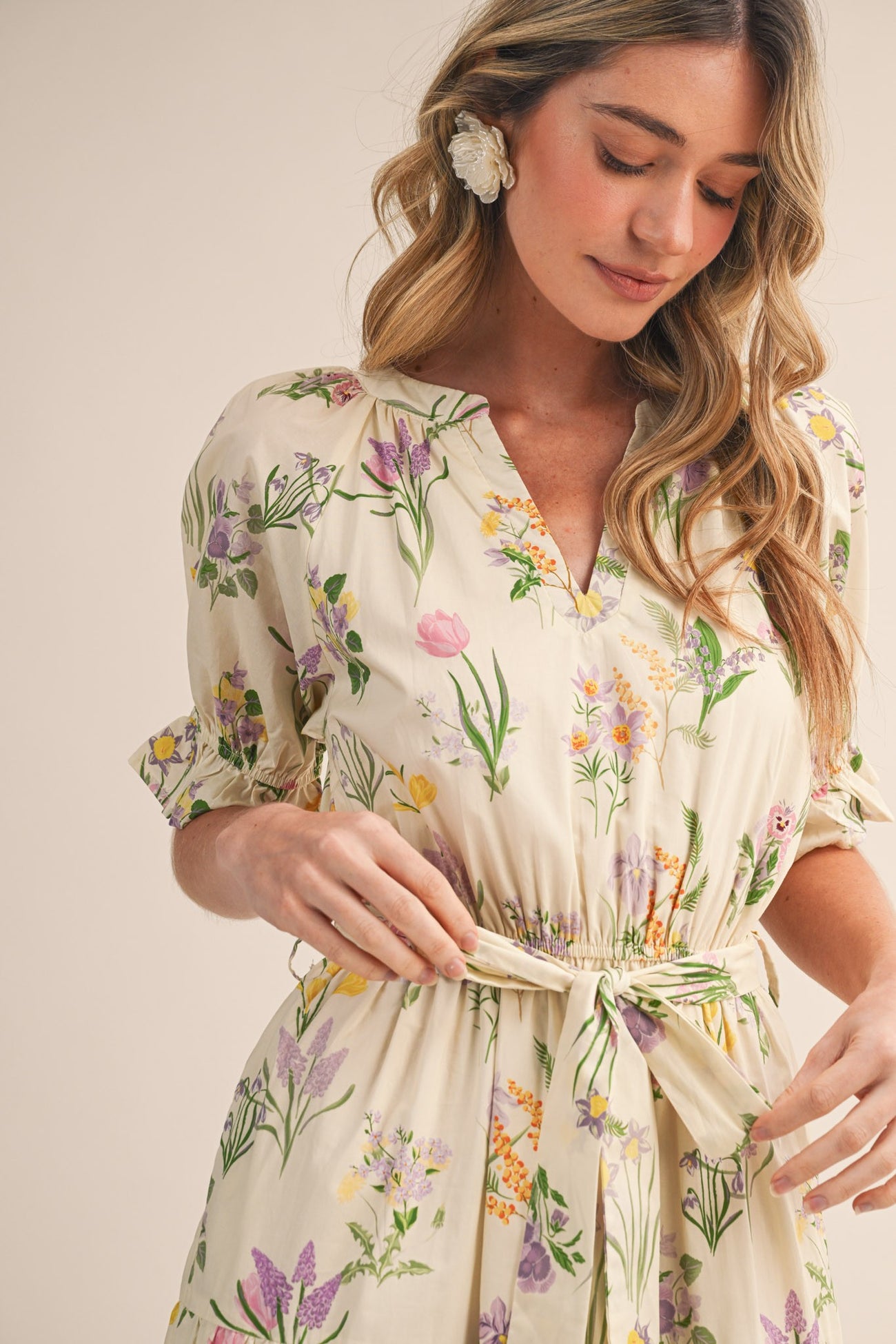 woman wearing a floral print cream dress with a v-neck and cinched waist, styled with floral earrings