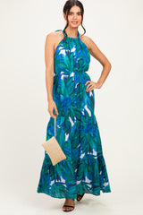 woman wearing a tropical print halter neck maxi dress in shades of blue and green, holding a straw bag