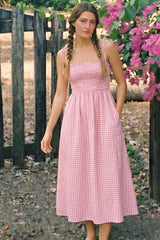 woman wearing a pink gingham smocked maxi dress with adjustable straps and pockets, standing outdoors among flowers