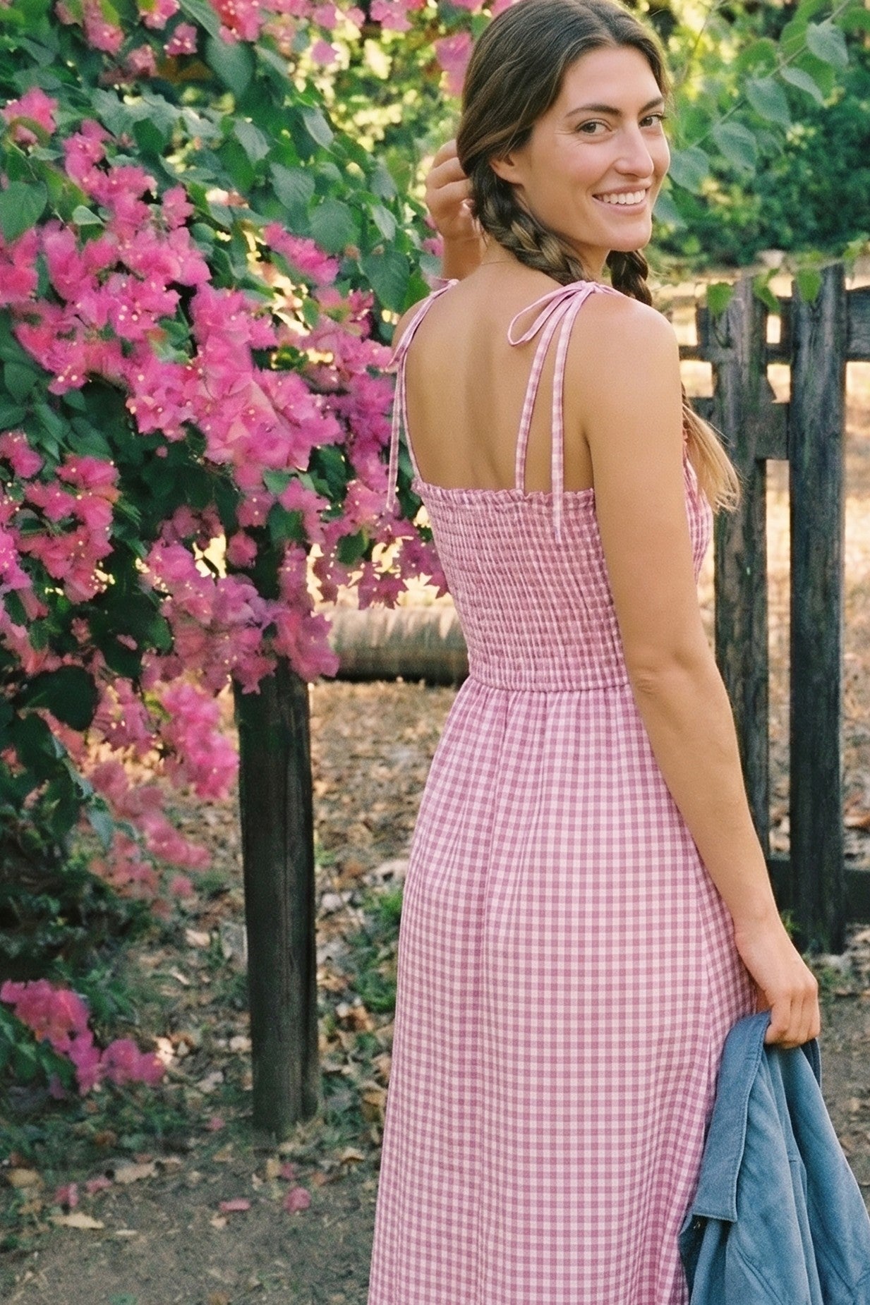 woman wearing a pink gingham midi dress with a fitted bodice and tie straps, standing outdoors by pink flowers