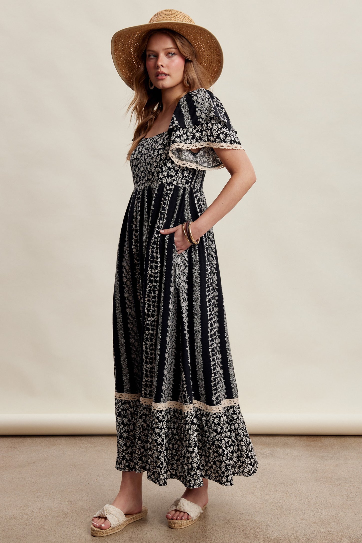woman wearing a black and white floral maxi dress with short ruffled sleeves and pockets, paired with a straw hat and sandals