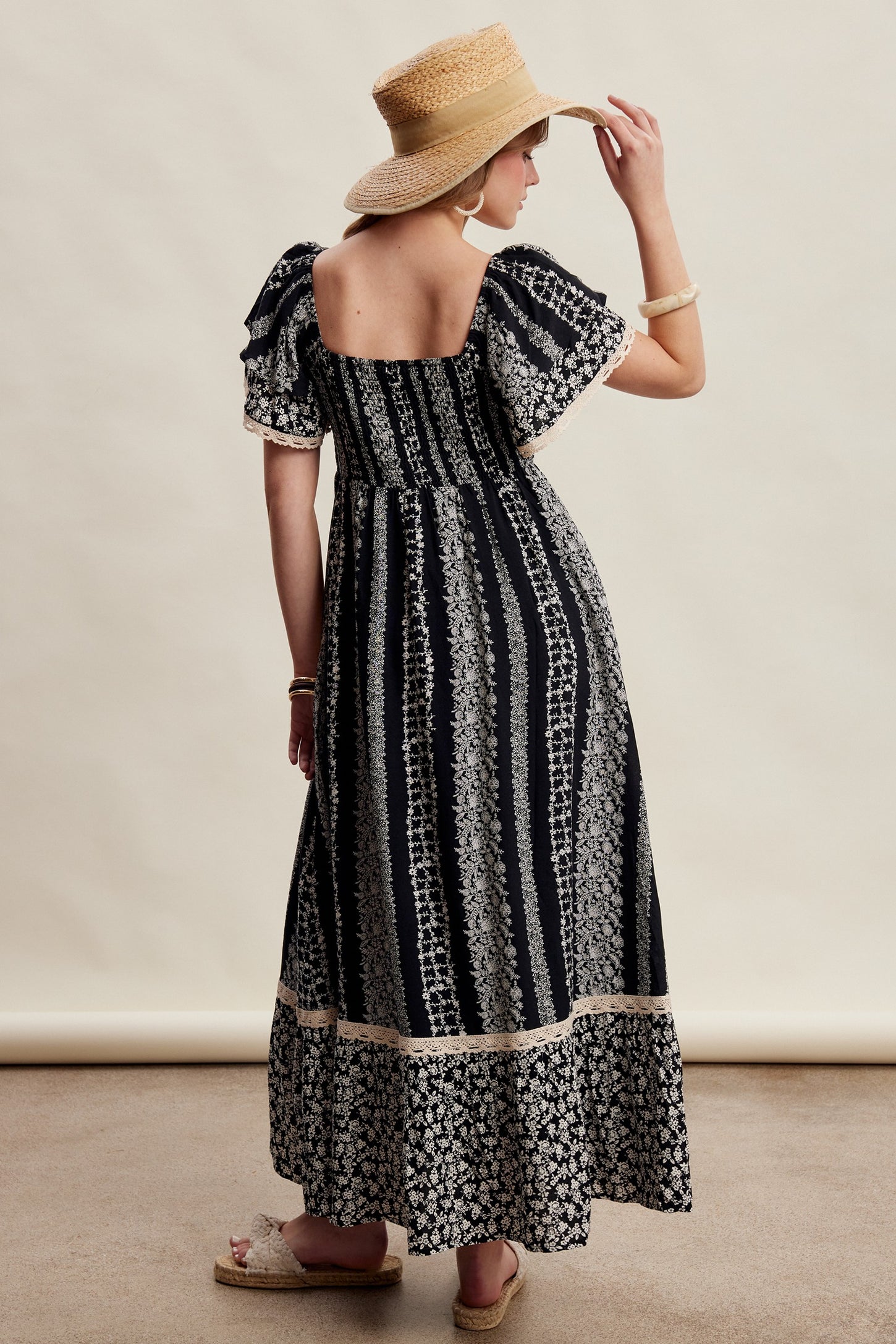 woman wearing a navy blue maxi dress with white floral patterns and puff sleeves, standing with a straw hat and sandals