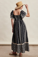 woman wearing a navy blue maxi dress with white floral patterns and puff sleeves, standing with a straw hat and sandals