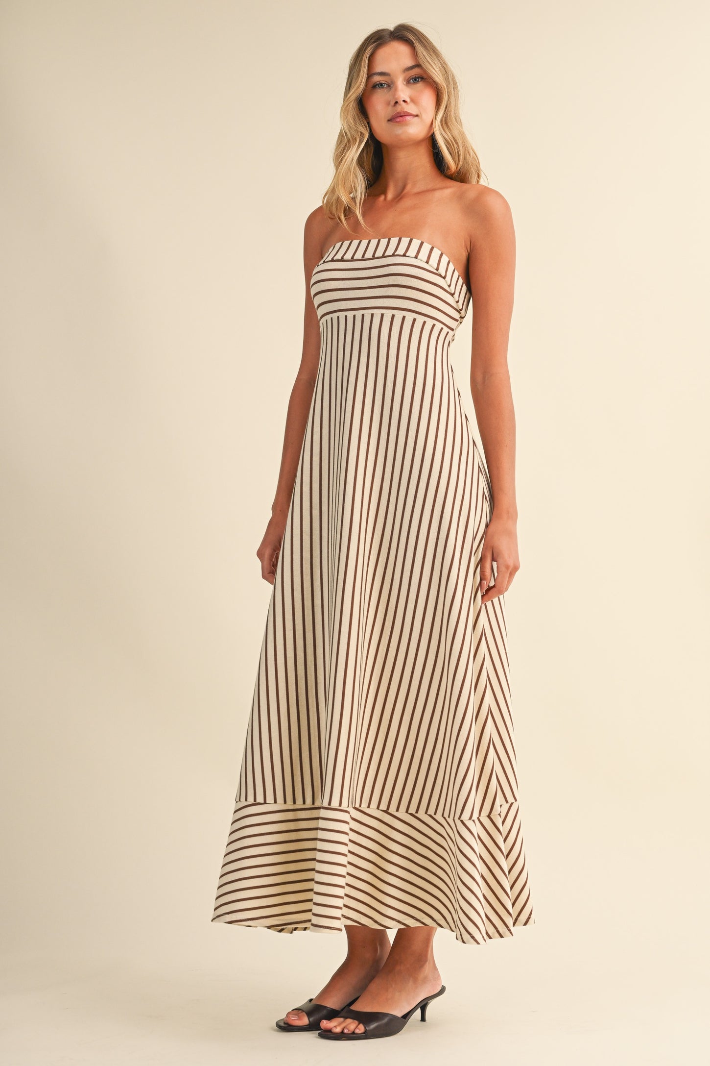 woman wearing a strapless beige maxi dress with vertical brown stripes and a flared hem, standing against a light background