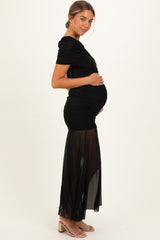 woman wearing a black maternity gown with ruched detailing and a sheer overlay, showing her baby bump