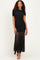 woman wearing a black ruched maxi dress with sheer overlay and short sleeves, standing against a neutral background