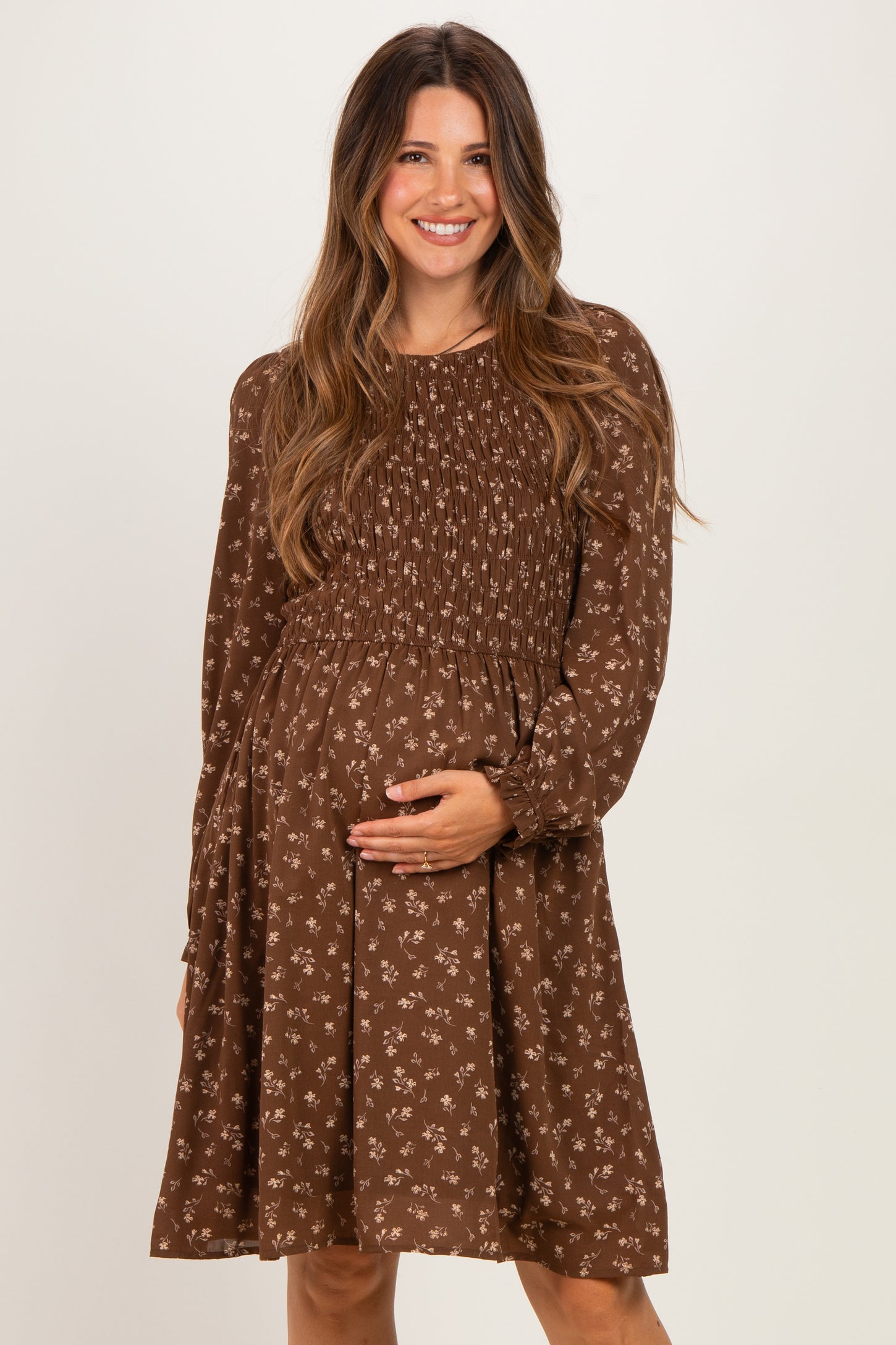 woman wearing a brown floral maternity dress with smocked bodice and long sleeves, holding her baby bump