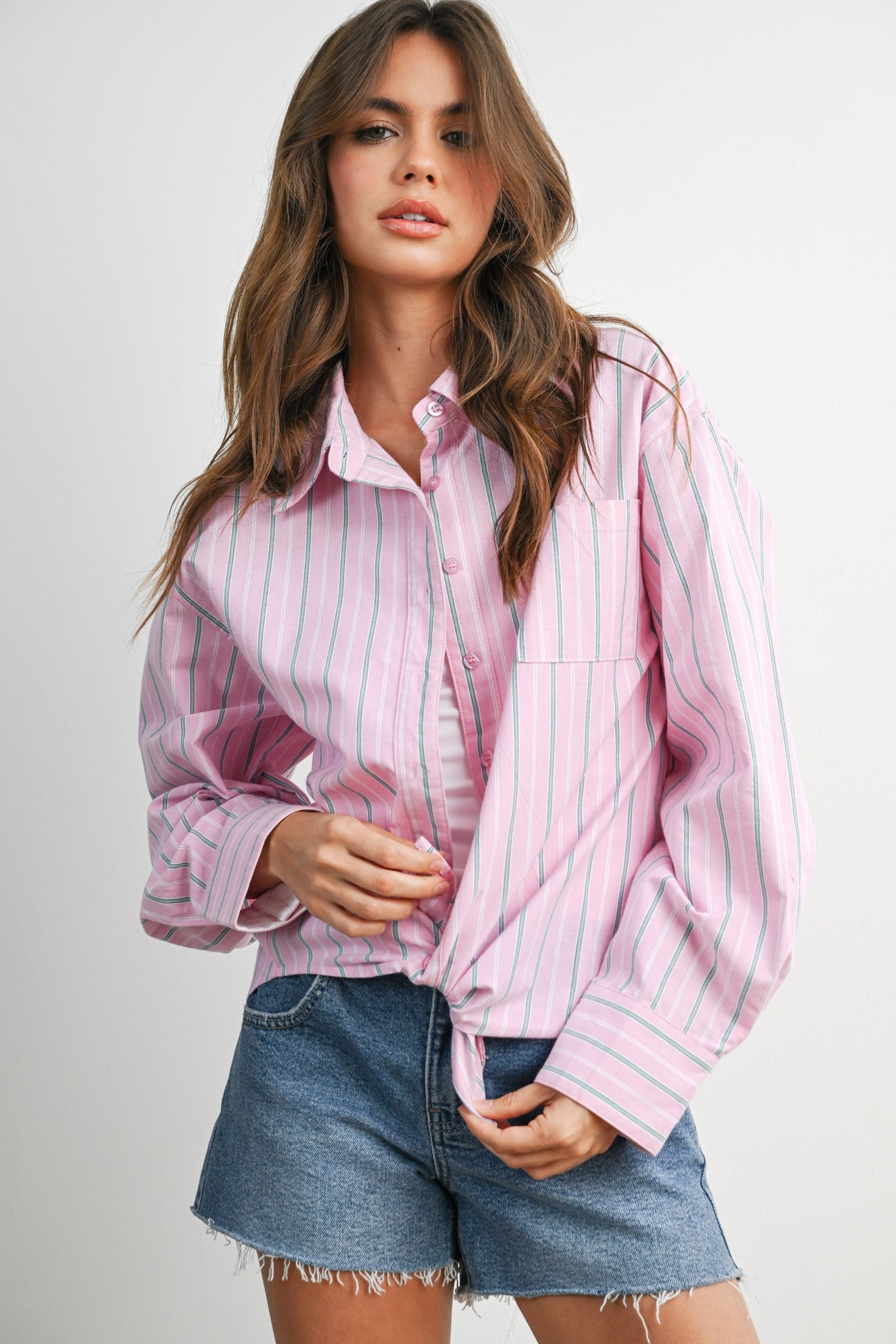 woman wearing a pink and white striped oversized button-up shirt knotted at the waist with denim shorts