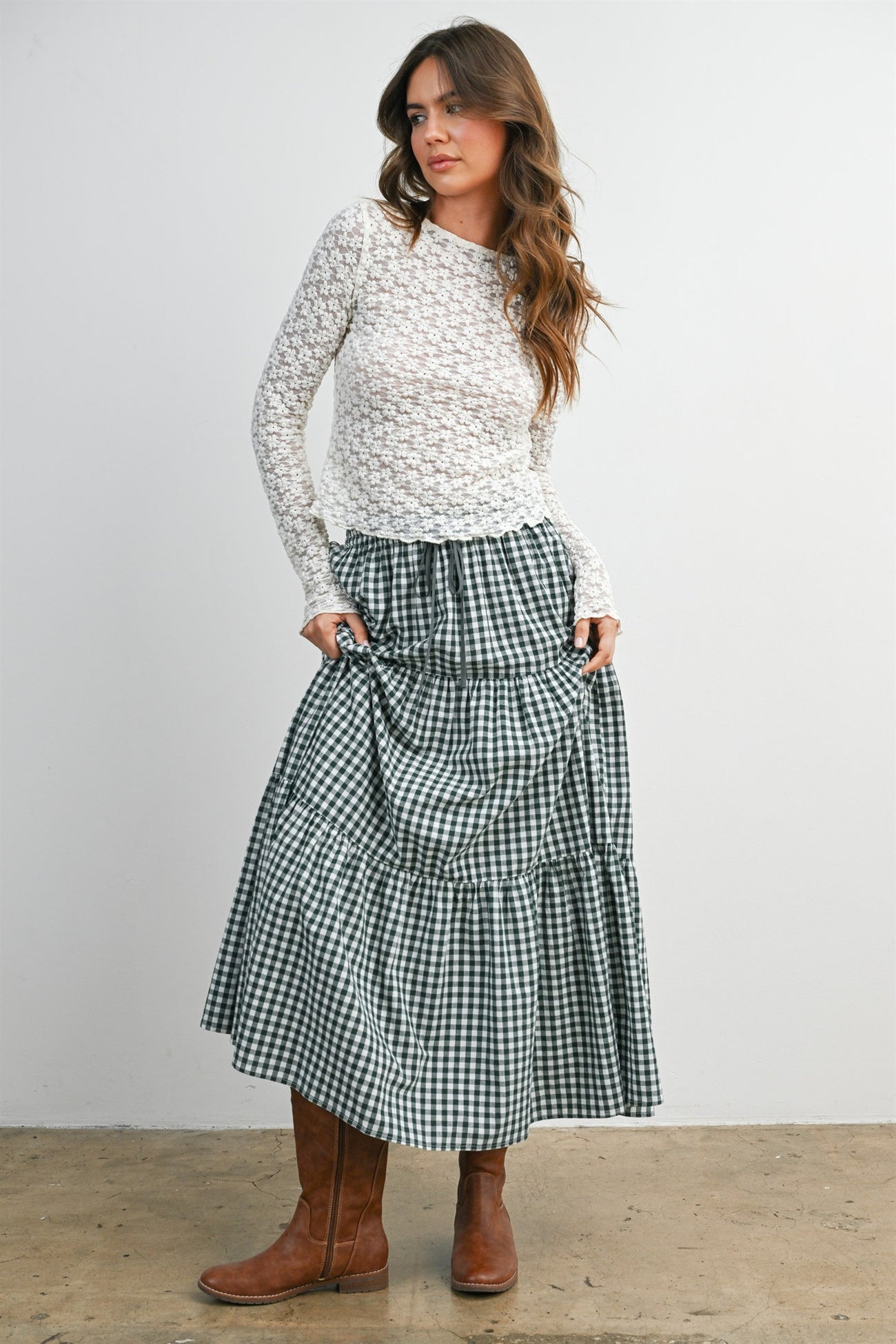 woman wearing a white lace long sleeve top and a green and white gingham tiered maxi skirt with brown knee-high boots