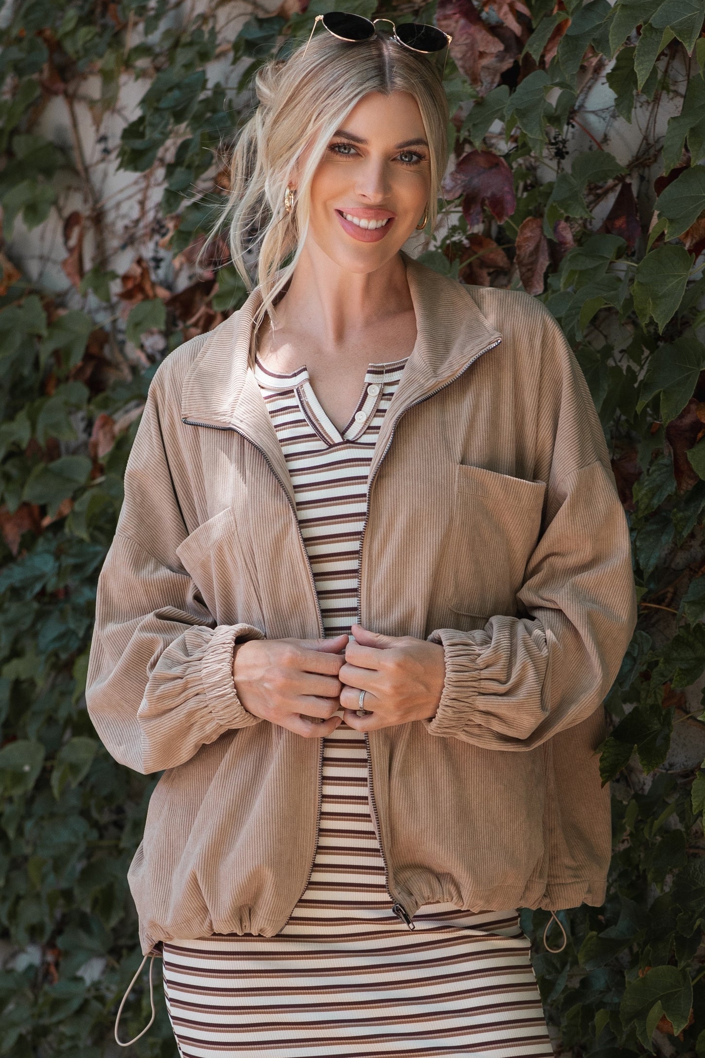 woman wearing a beige corduroy zip-up jacket over a striped dress, smiling against a leafy background