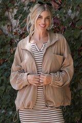 woman wearing a beige corduroy zip-up jacket over a striped dress, smiling against a leafy background