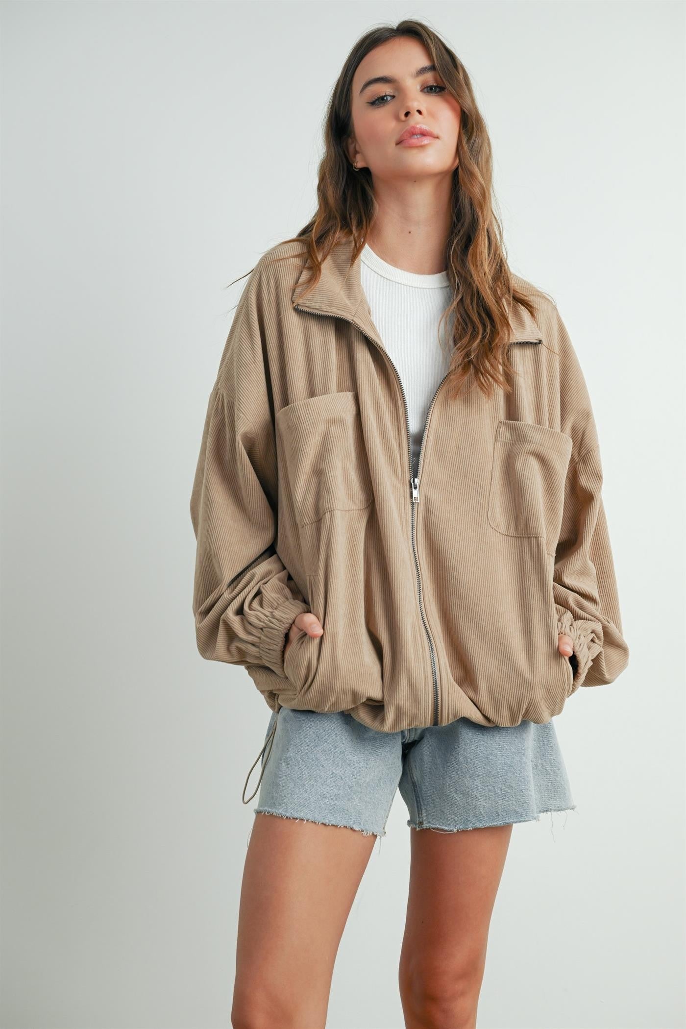 woman wearing a beige zip-up jacket with two front pockets and elastic cuffs paired with light blue denim shorts