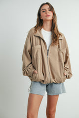 woman wearing a beige zip-up jacket with two front pockets and elastic cuffs paired with light blue denim shorts