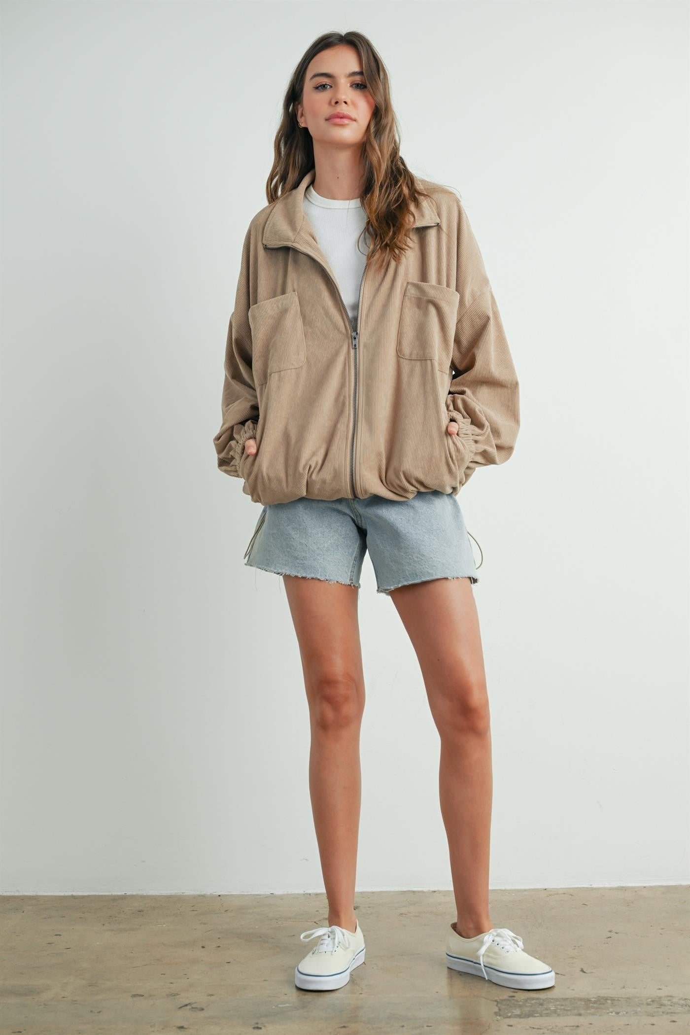 woman wearing a beige zip-up corduroy jacket, white t-shirt, and light denim shorts with white sneakers